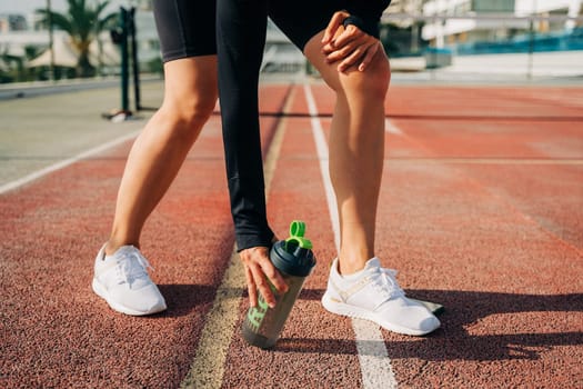 Close up of woman runner legs, standing on road and relaxing after sport training. Holding water blender bottle while doing workout in summer city street.
