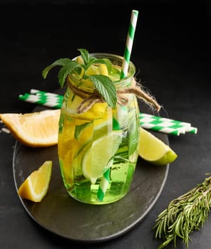 Lemonade in a transparent glass with lemon, lime, rosemary sprigs and mint leaves on a black background