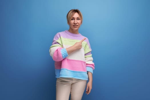 pleasant blond woman in casual outfit shows her hand to the side on a blue background with copy space.