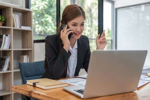 Cheerful young beautiful business woman talking on mobile phone and using laptop with smile while sitting at her working place.