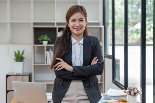 Young successful woman entrepreneur or an office worker stands with crossed arms near a desk in a modern office, looking at the camera and smiling.