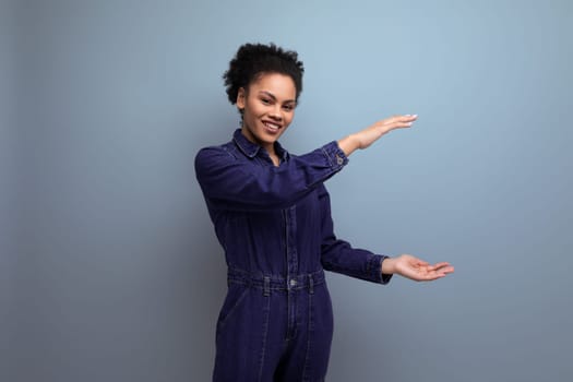 young hispanic business woman with fluffy hair dressed in blue denim overalls shows with her hands the size of an object.