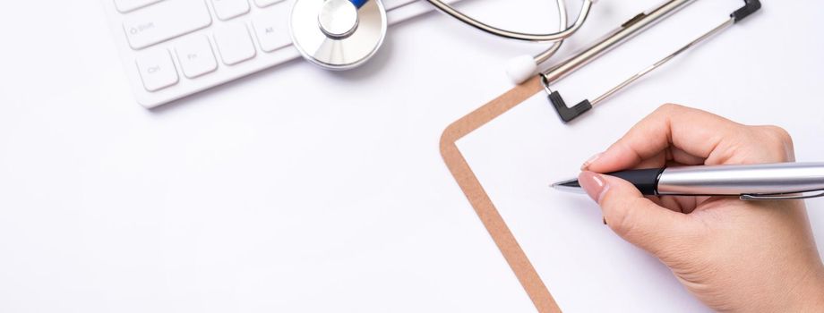 Female doctor writing a medical record case over clipboard on white working table with stethoscope, computer keyboard. Top view, flat lay, copy space