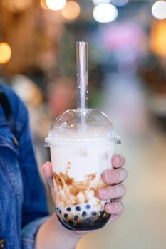 Young girl in denim jacket is drinking brown sugar flavored tapioca pearl bubble milk tea with glass straw in night market of Taiwan, close up, bokeh