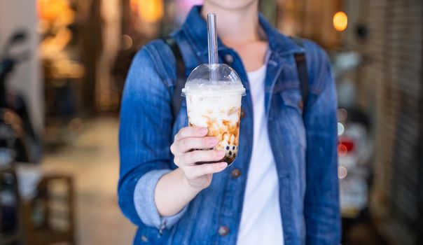 Young woman is holding, drinking brown sugar flavored tapioca pearl bubble milk tea with glass straw in night market of Taiwan, close up, bokeh