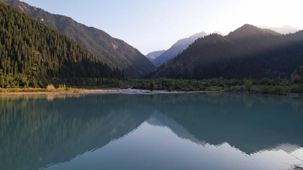 View of the mountain lake Issyk from a height. Turquoise-emerald water from the glacier. Reflection of mountains, forests and the sun on the water. The river flows into the lake. Issyk Dam. Kazakhstan