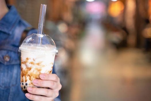 Young girl in denim jacket is drinking brown sugar flavored tapioca pearl bubble milk tea with glass straw in night market of Taiwan, close up, bokeh