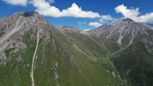 Clouds flow around the tops of high mountains. Rocky gorges and cliffs. The sky is blue, the sun is shining brightly, green grass is growing in places. Mountain terrain from the height of the drone