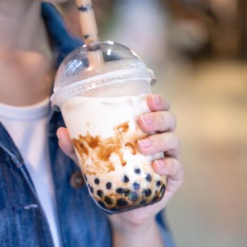 Young woman is holding, drinking brown sugar flavored tapioca pearl bubble milk tea with glass straw in night market of Taiwan, close up, bokeh