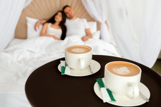 Waiter delivering 2 cups of coffee to a hotel room for young married couple lying in a bed