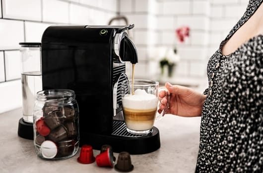 Girl with capsule coffee machine and cappuccino in transparent cup at kitchen. Woman preparing italian caffeine beverage using professional espresso maker