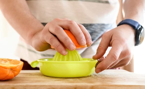 Man hand squeeze ripe orange with manual extractor for making natural juice at kitchen. Guy preparing tropical fruit drink from citrus at home