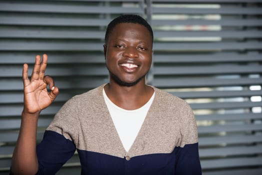 young man standing in a room staring at the camera smiling while making fingers.