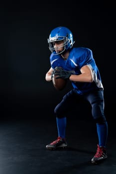 Full length portrait of a man in a blue american football uniform against a black background. Sportsman in a helmet with a ball