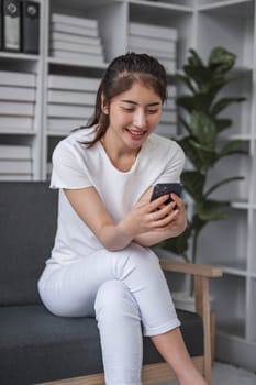 Attractive and happy young Asian female using her smartphone to scroll through social media while relaxing on her comfortable sofa in her living room. ..