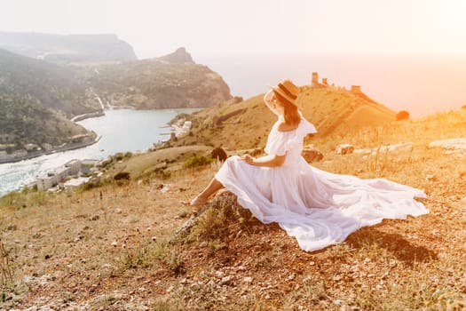 Happy woman in a white dress and hat stands on a rocky cliff above the sea, with the beautiful silhouette of hills in thick fog in the background