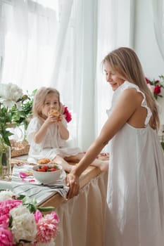 A little blonde girl with her mom on a kitchen countertop decorated with peonies. The concept of the relationship between mother and daughter. Spring atmosphere