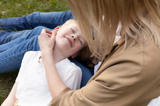 Cute Caucasian 7 yo blonde boy lying on parents knees in park. Mom stroking son's smiling face. Summer time. Parenthood, family leisure time. Love and Care. Closeup Horizontal Plane High quality photo