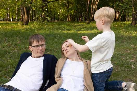 Cute Caucasian 7 Yo Boy Puts, Inserts Flower In Mother's Hair In Park. Mom And Dad Lying On Grass. Summer Time. Parenthood, Family Leisure Time. Love And Care. Horizontal Plane. High quality photo