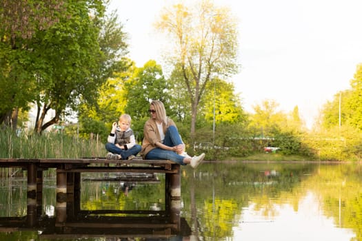 Caucasian Mother And Son Sit On Wooden Dock Enjoying Lake View In Summer Time. Boy Throws Rocks Into Water. Mom's Support And Care. Motherhood, Family Leisure Time. Children's Day. Horizontal Plane.
