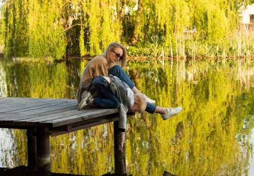 White Mother And Son Sit On Wooden Dock Enjoying Lake View In Summer Time. Child Throws Rocks Into Water. Mom's Support And Care. Motherhood, Family Leisure Time. Children's Day. Horizontal Plane.