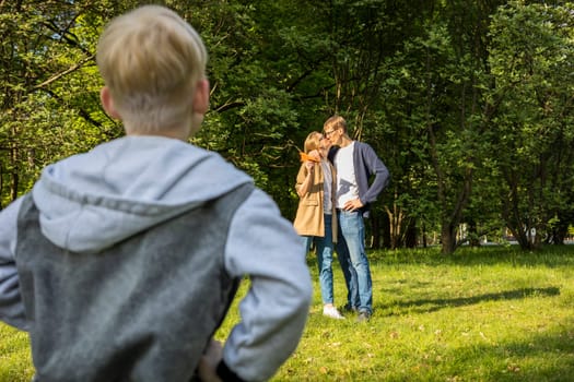 Back View OF Emotional Caucasian Boy Looking At Kissing Parents in Park. Kid's Jealousy. Mom Hugs Dad. Parenthood, Child' Feeling. Horizontal Plane. High quality photo