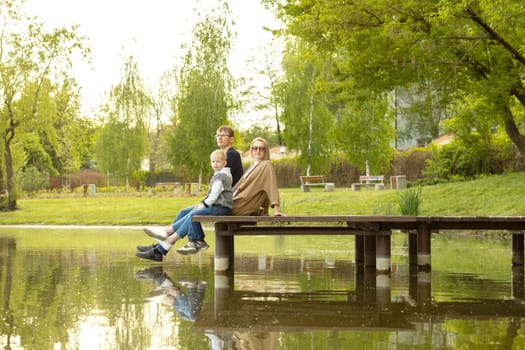Caucasian Family Mother, Father And Son Sit On Wooden Dock Enjoying Lake View In Summer Time. Family Support And Care. Parenthood, Family Leisure Time. Children's Day. Horizontal Plane.
