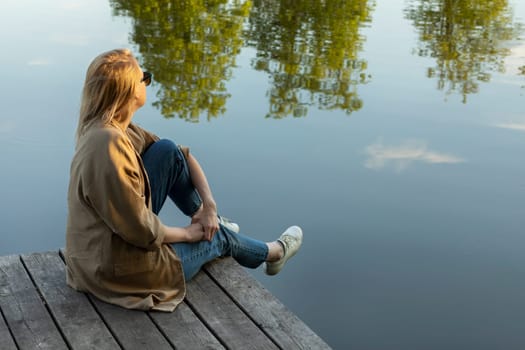 Young Pretty Blonde Woman Sit On Wooden Dock Enjoying Lake View In Summer Time on Sunset. Rest And Recharge in Nature, Self Awareness, Leisure Time, Mental Health. CopySpace For Text Horizontal Plane