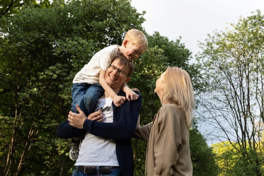 Father Carried Little Child, Son On Shoulder. Mother Walks Beside In Park. Summer Time. Happy Parenthood, Family Leisure Time. Love And Care. Horizontal Plane. High quality photo