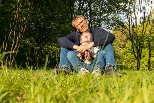 Father And Child, Son Sitting On Grass In Park, Enjoying Time Together. Summer Time. Happy Parenthood, Family Leisure Time, Father's Day. Emotional Connection, Love And Care. Horizontal Plane.