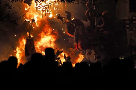 Horizontal photo of people watch Ninote dolls go up in flames in a street lit by lights during the Fallas celebrations in Valencia, Spain