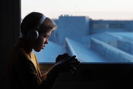 Horizontal photo of a 12-year-old teenager sits by the window overlooking the city, wearing white headphones and looking at their phone