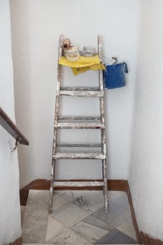 Vertical photo of a repair kit consisting of a bucket, a roller, a brush, tape, and a folding ladder on the landing in the entrance of the building