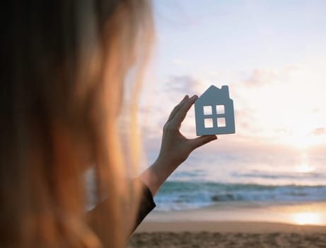 Horizontal photo of a woman is holding a figurine of a white house, against the backdrop of the sky with clouds and the sea during sunset