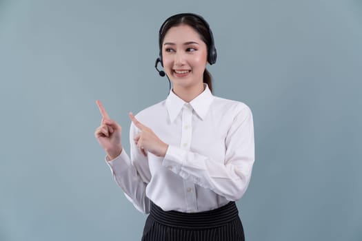 Asian female call center operator with smile face advertises job opportunity, wearing a formal suit and headset pointing finger for product on customizable isolated background. Enthusiastic