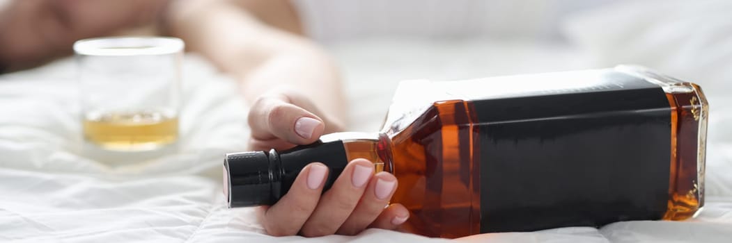 Woman is lying in bed dead drunk holding almost empty bottle of booze. Intoxicated woman after hard night party.