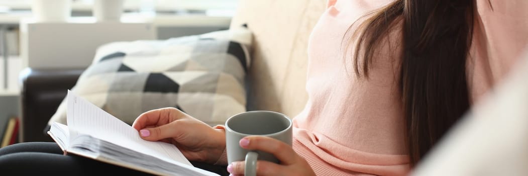 Smiling woman is reading a book and holding cup of tea on sofa. Home leisure and hobby concept