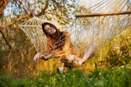 a joyful woman with long hair is lying in a hammock in an orange dress and happily smiling at the camera. A photo taken on the street on the theme of recreation in the country. High quality photo
