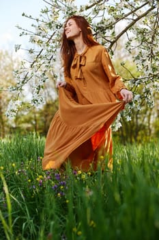 a happy, slender, sweet woman stands in a long orange dress in the tall grass near a flowering tree and happily smiling lifts the hem of her dress. High quality photo