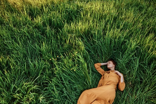 a sweet, calm woman in an orange dress lies in a green field enjoying the silence and peace. Horizontal photo taken from above. High quality photo