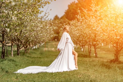 Blond blooming garden. A woman in a white dress walks through a blossoming cherry orchard. Long dress flies to the sides