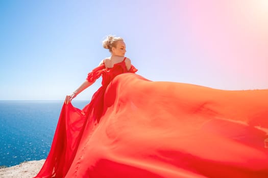 woman sea red dress. Blonde with long hair on a sunny seashore in a red flowing dress, back view, silk fabric waving in the wind. Against the backdrop of the blue sky and mountains on the seashore