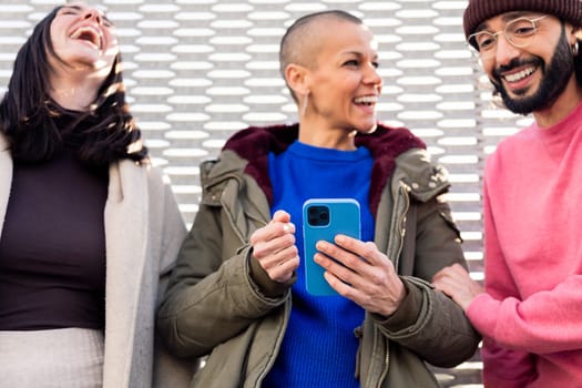 three young friends laughing and enjoying the moment using together a mobile phone, focus on the phone, concept of technology of communication and modern lifestyle