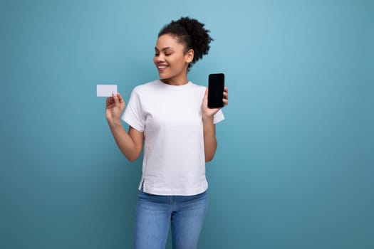 calm young brunette hispanic woman in a white t-shirt shows a mockup of a credit card and a smartphone on the background with copy space.