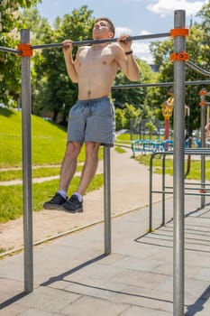 Athletic caucasian topless muscular man doing pull ups exercises on horizontal bar. Young guy pumping up back muscles on summer playground. Sports health fitness routine, workout. Strength, motivation