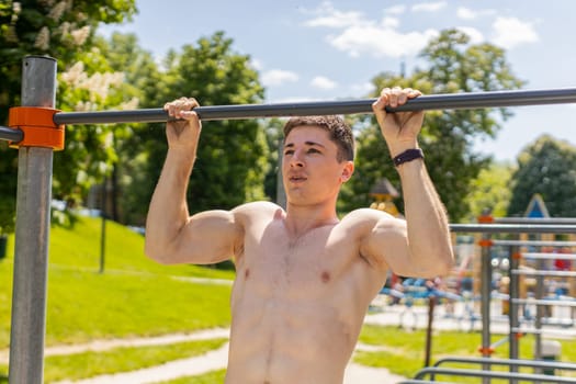 Athletic caucasian topless muscular man doing pull ups exercises on horizontal bar. Young guy pumping up back muscles on summer playground. Sports health fitness routine, workout. Strength, motivation