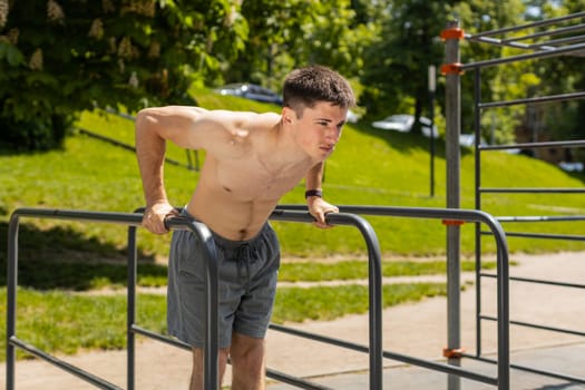 Athletic caucasian topless muscular man practicing dips on parallel bars, training triceps. Young shirtless guy on playground. Sports health fitness routine, workout. Strength motivation. Outdoor gym