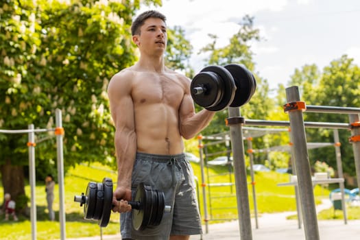 Athletic caucasian topless muscular man doing arms weight lifting dumbbells exercises, pumping up arm bicep muscles. Young guy on playground. Sports health fitness routine workout. Strength motivation