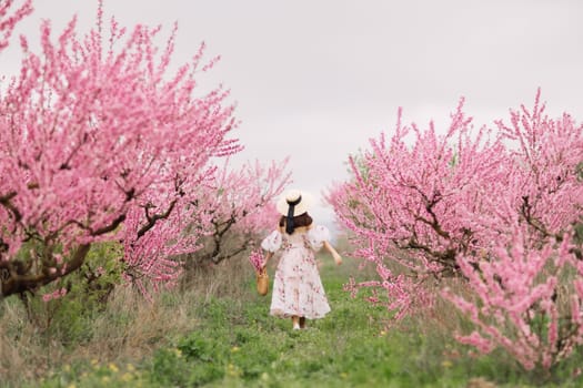 Woman blooming peach orchard. Against the backdrop of a picturesque peach orchard, a woman in a long dress and hat enjoys a peaceful walk in the park, surrounded by the beauty of nature