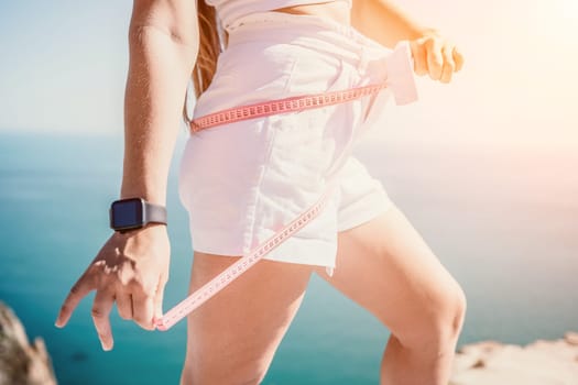 a young and slender woman in shorts holds in her hand a measuring yellow tape on her sexy thighs on the beach. concept of detox and diet